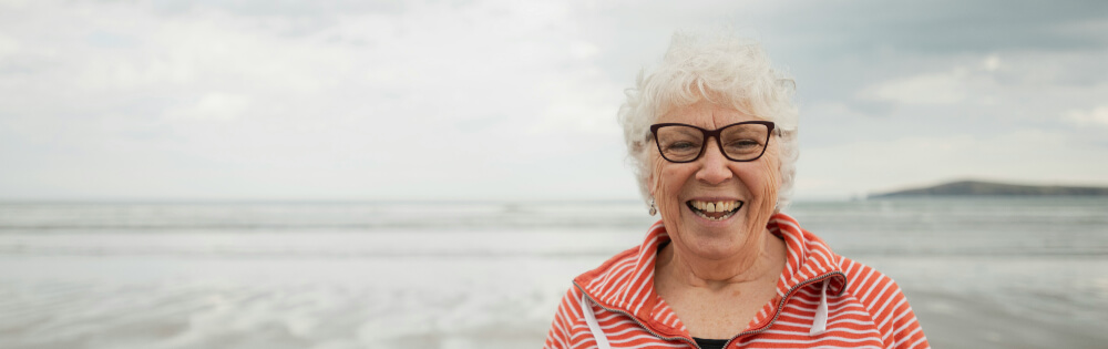 Sterbegeldversicherung Eine ältere Frau mit weißem Haar und Brille lächelt fröhlich am Strand. Sie trägt ein gestreiftes Oberteil und im Hintergrund ist das Meer mit einem wolkigen Himmel zu sehen.